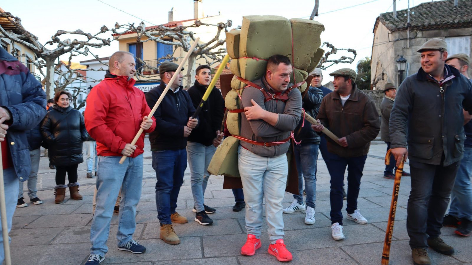 Un hombre con pantalones claros sostiene en su espalda bloques de espuma verde atados, rodeado por varias personas en una plaza.