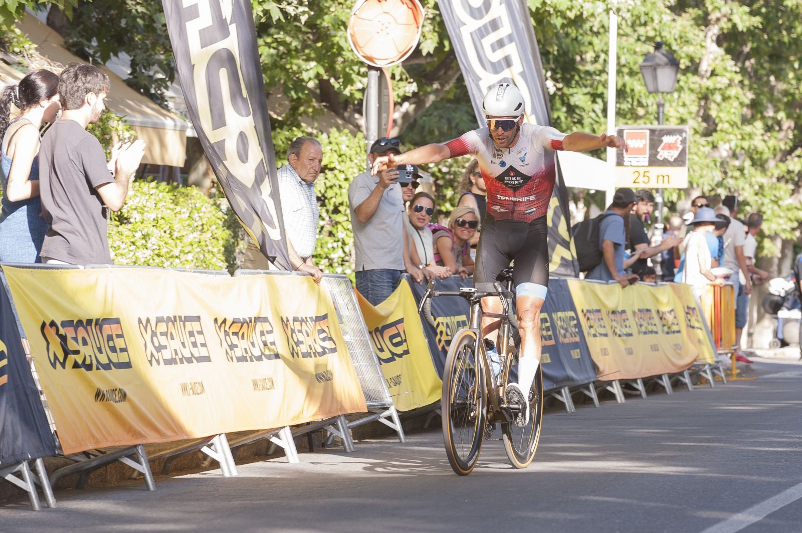 Un ciclista con los brazos extendidos cruza la línea de meta rodeado de vallas publicitarias y público aplaudiendo.