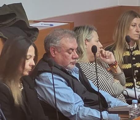 Plano medio de varias personas sentadas en bancadas de madera en una sala de plenos municipal con micrófonos individuales.