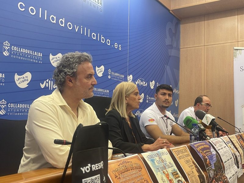 Cuatro personas sentadas de perfil en una mesa con micrófonos y carteles informativos sobre un fondo azul.