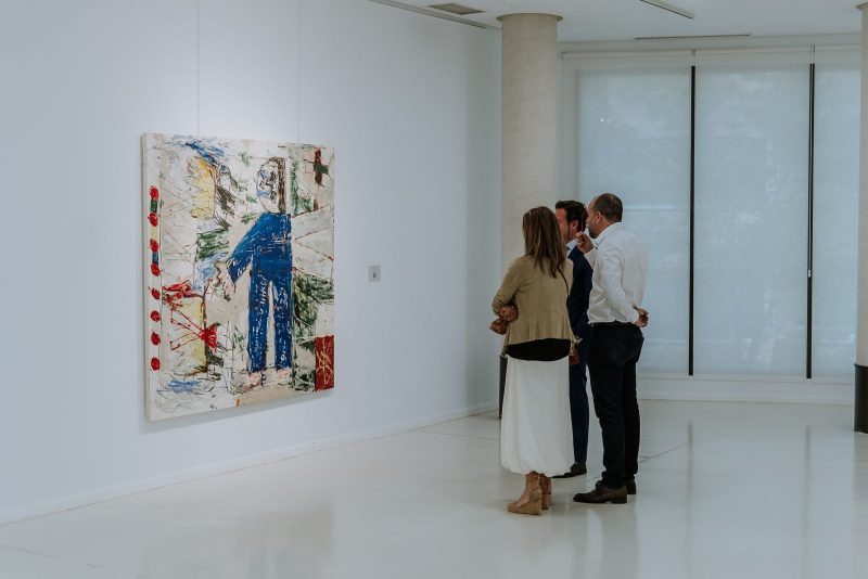 Tres personas de espaldas contemplan una gran pintura de estilo expresionista colgada en una pared blanca de una galería.