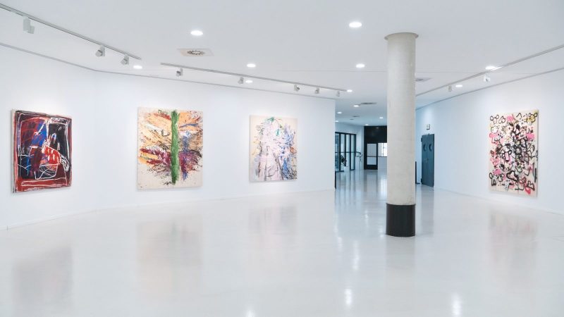 Amplia sala de arte iluminada, de paredes y suelos blancos, con varios cuadros abstractos de gran formato colgados en hilera.