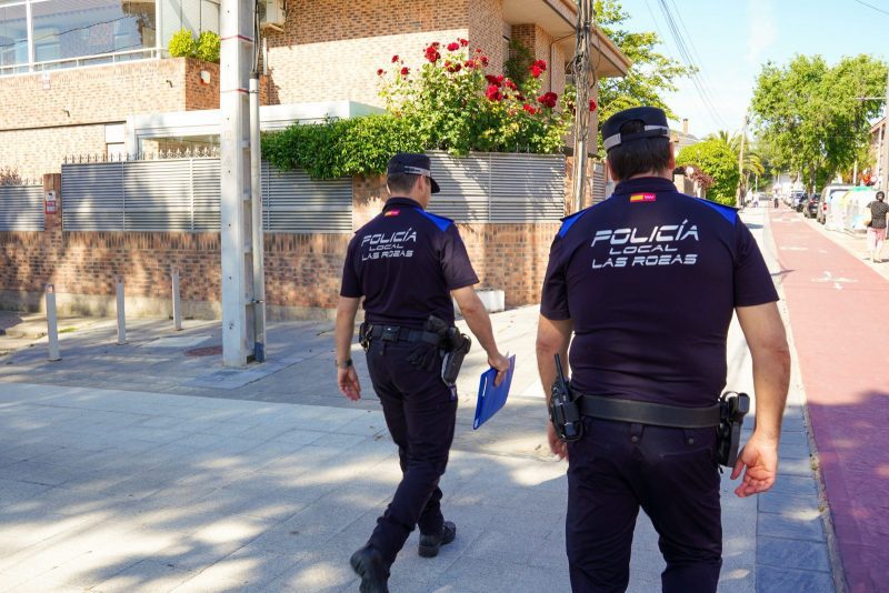 Dos agentes de la Policía Local de Las Rozas uniformados patrullan a pie de espaldas por una acera residencial.