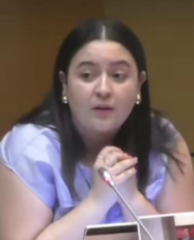 Plano medio de una mujer con el cabello oscuro hablando frente a un micrófono con las manos juntas.