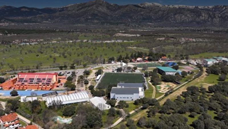 Vista aérea de un complejo deportivo municipal con campos de fútbol, pistas y montañas al fondo bajo un cielo despejado.