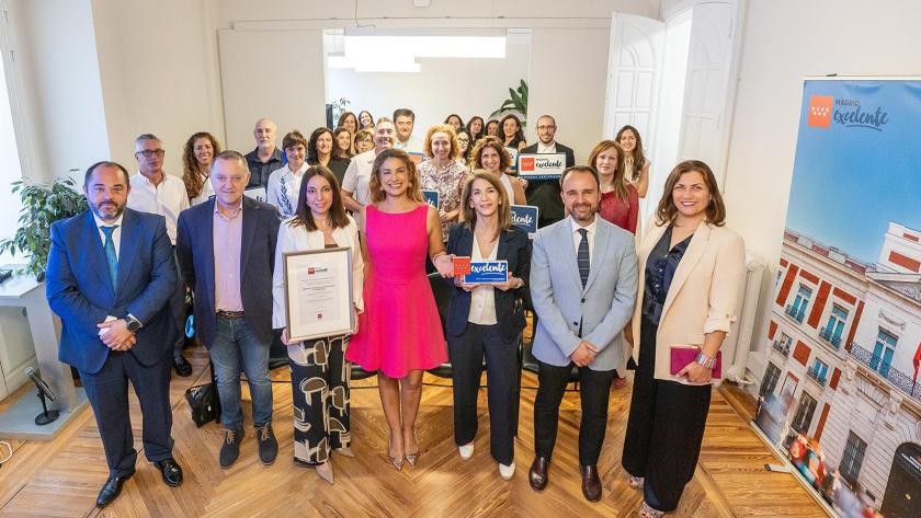 Un grupo numeroso de personas posa en una sala con suelo de madera, sosteniendo un certificado enmarcado y un distintivo azul.