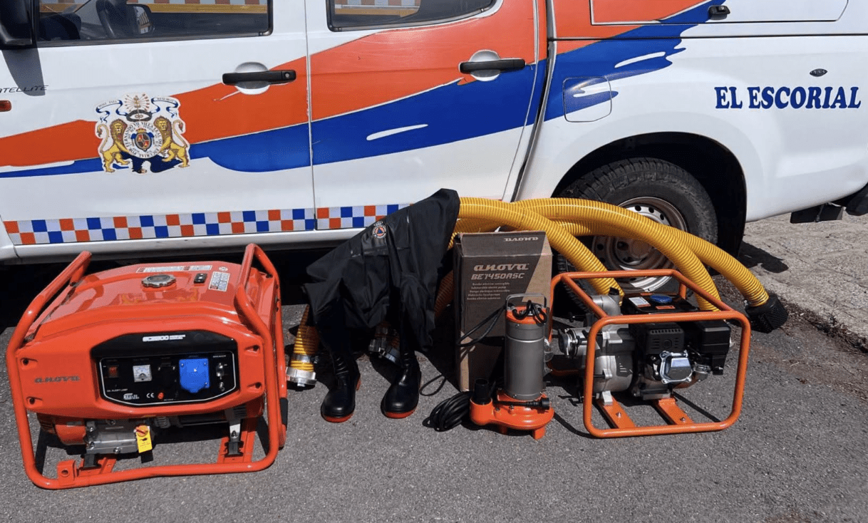 Un generador eléctrico naranja, una motobomba, mangueras amarillas y equipamiento técnico expuestos junto a un vehículo de emergencias de El Escorial.