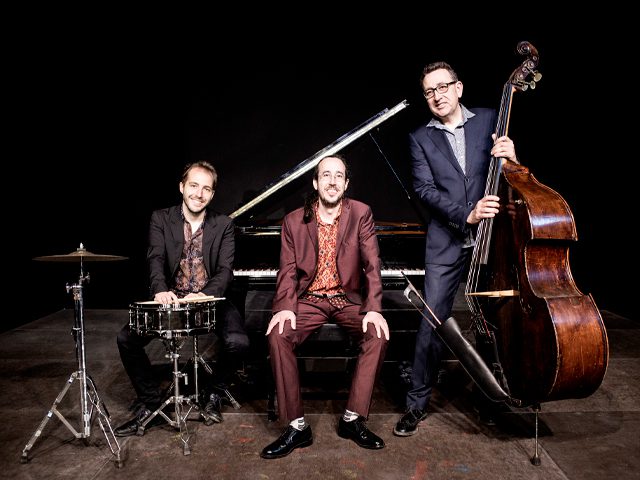 Tres músicos sonriendo posan junto a un piano de cola, un contrabajo y una caja de batería sobre fondo negro.