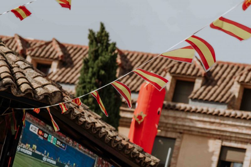 Banderines con la bandera de España cuelgan frente a tejados rústicos y una pantalla gigante de televisión.