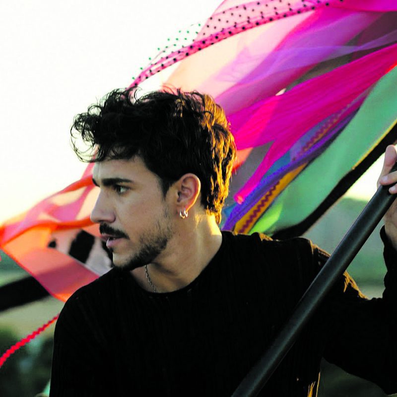 Retrato de perfil del cantante Nil Moliner luciendo un pendiente de aro y barba corta, con telas de colores al fondo.