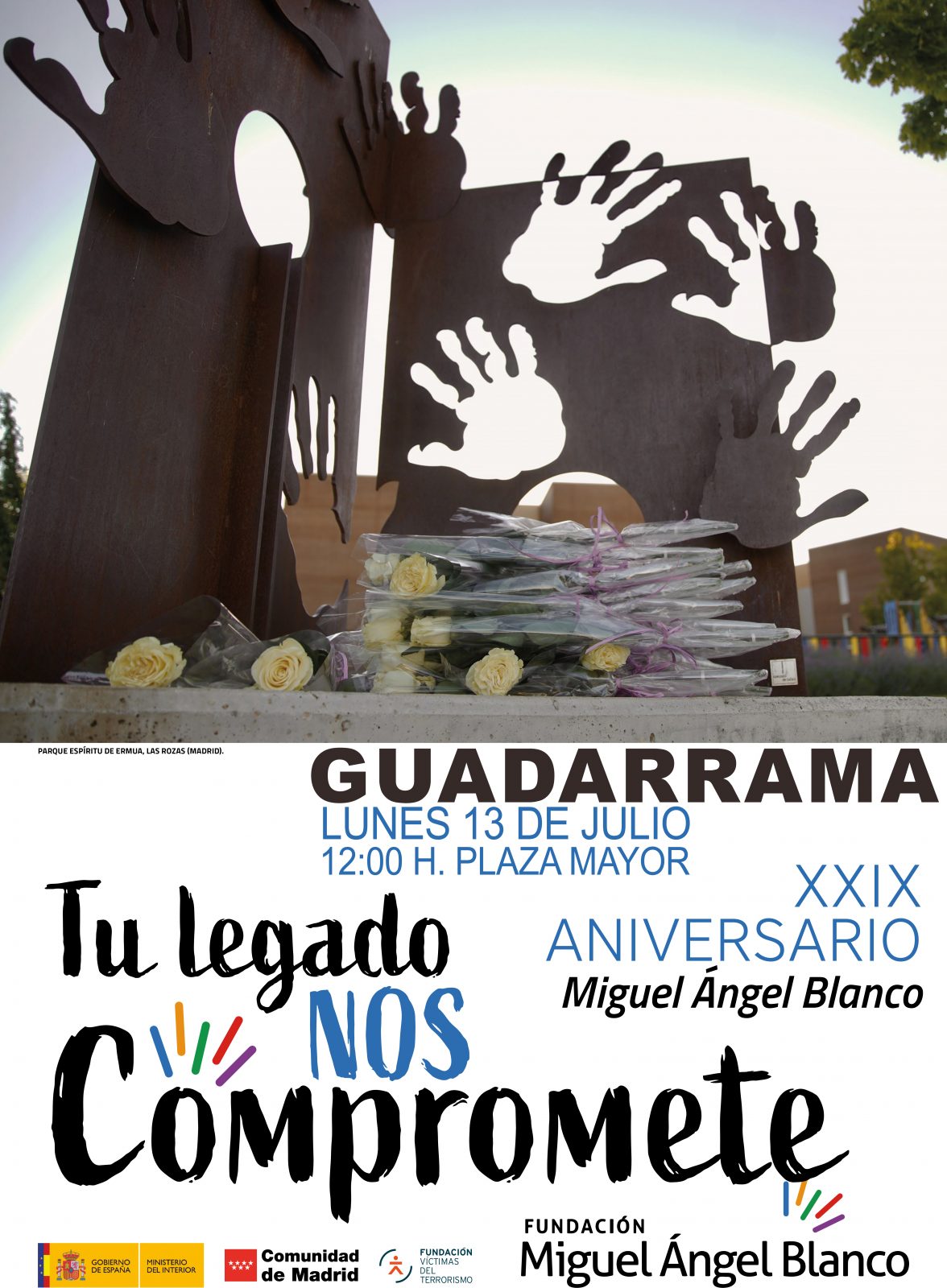Monumento metálico con siluetas de manos recortadas que tiene flores blancas en la base, sobre un cartel informativo de un acto.