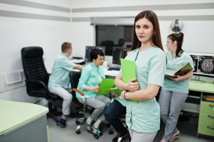 Una joven profesional de la salud sostiene una carpeta verde en un laboratorio con sus compañeros al fondo.