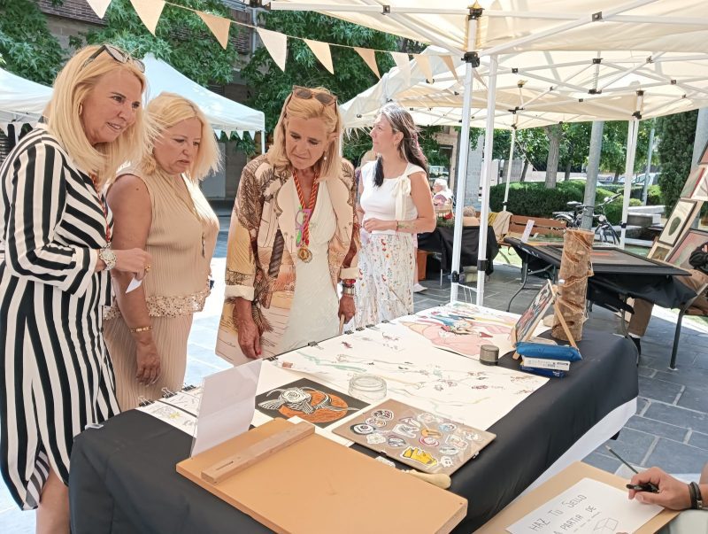 La alcaldesa de Collado Villalba, Mariola Vargas, y varias asistentes observan obras en un puesto de la feria.