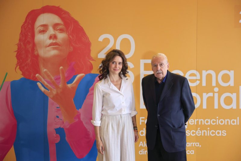 La presidenta Isabel Díaz Ayuso posa junto a un hombre con traje oscuro ante un cartel amarillo.