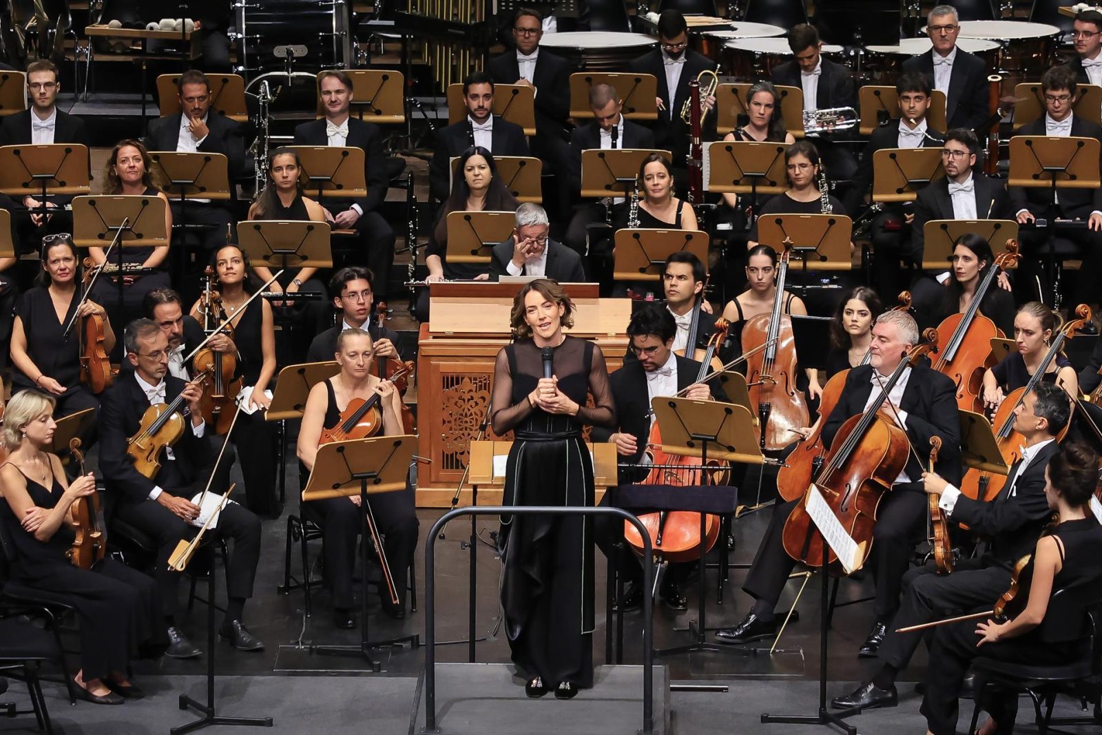 La soprano Raquel Lojendio habla con un micrófono sobre el escenario frente a la orquesta.