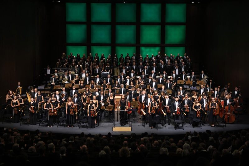 Una gran orquesta y coro actúan sobre un escenario iluminado frente al público asistente.