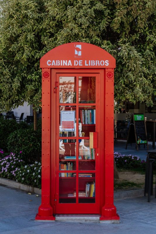 Vista frontal de una cabina roja con la puerta cerrada y estantes repletos de libros en su interior.