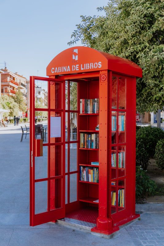 Cabina telefónica roja abierta llena de libros en una calle peatonal de Las Rozas.