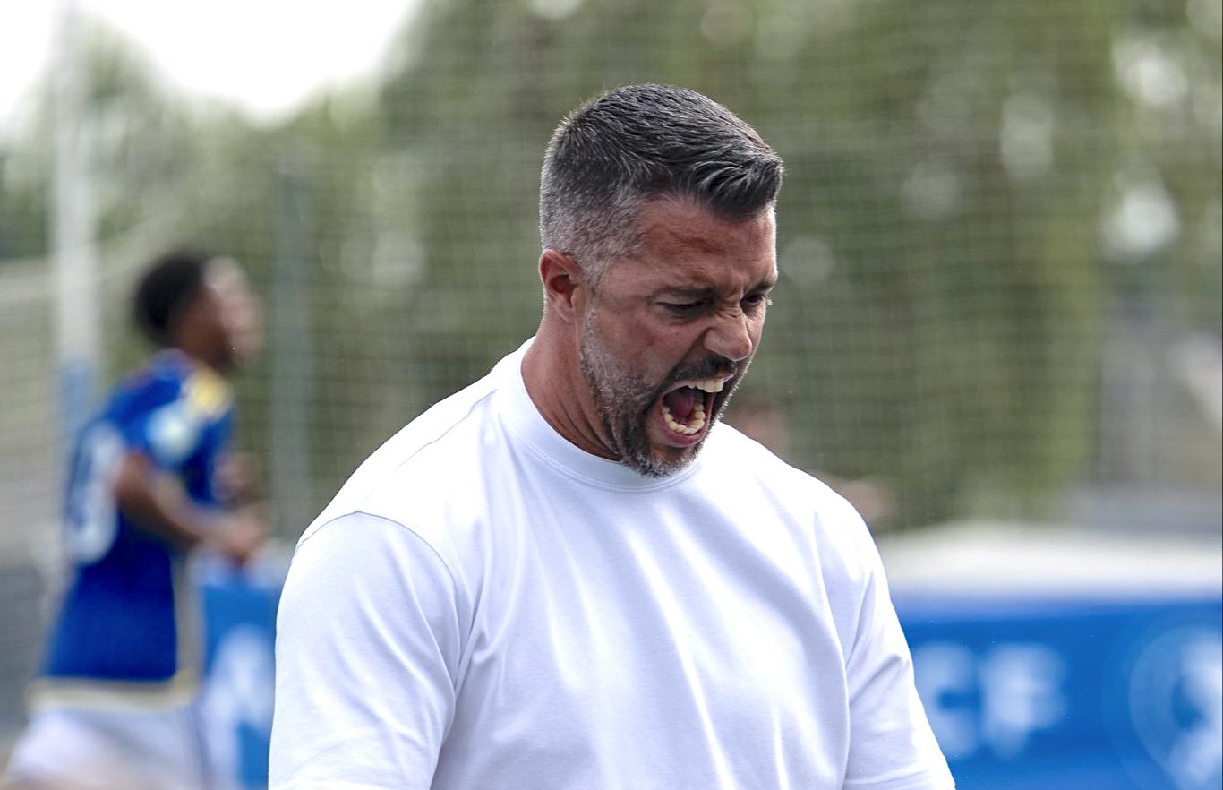 Entrenador gesticulando con intensidad durante un encuentro de fútbol en un campo al aire libre.