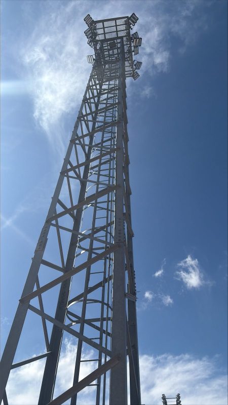 Torre metálica de iluminación de gran altura para un campo de fútbol bajo un cielo azul despejado.