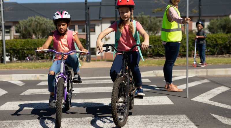 Side view of a blonde Caucasian woman wearing a high visibility vest and holding a stop sign, standing in the road on a pedestrian crossing and stopping the traffic while a Caucasian and an African American schoolgirl ride their bicycles across the road safely on their way to elementary school