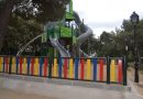 Parque infantil vallado con tablones de colores que rodea una torre de juegos con toboganes en un entorno arbolado.